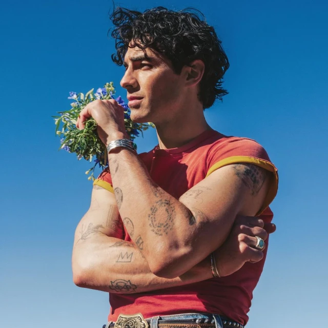 Man in a red t-shirt and ornate belt holds a bunch of blue flowers outdoors against a clear blue sky, portrait orientation, medium shot.