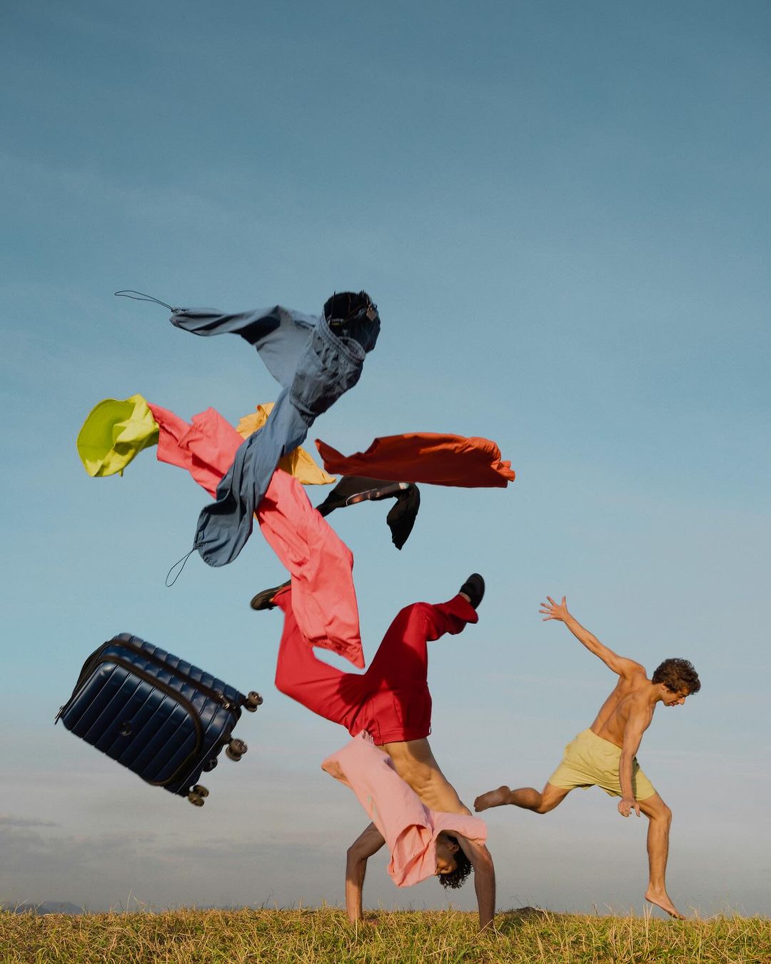 Two adults in outdoor setting with clothes suspended in mid-air and a suitcase, under a clear sky, showcasing a playful fashion scene.
