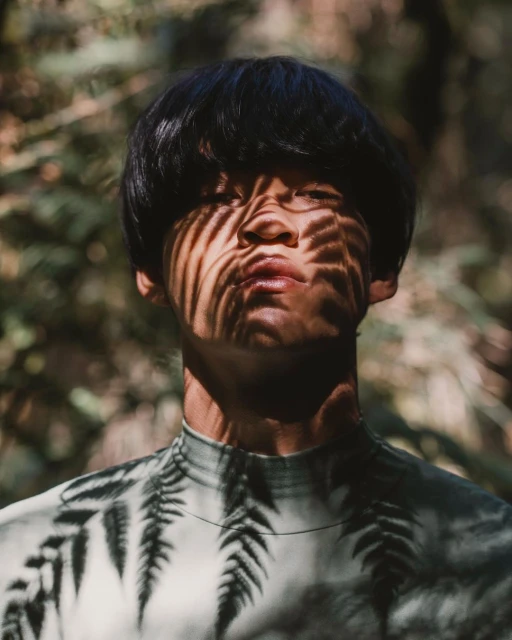 Portrait of an adult with dark hair in a fern-printed shirt, posing outdoors with shadows cast on their face from the sunlight filtering through trees.