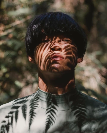 Portrait of an adult with dark hair in a fern-printed shirt, posing outdoors with shadows cast on their face from the sunlight filtering through trees.