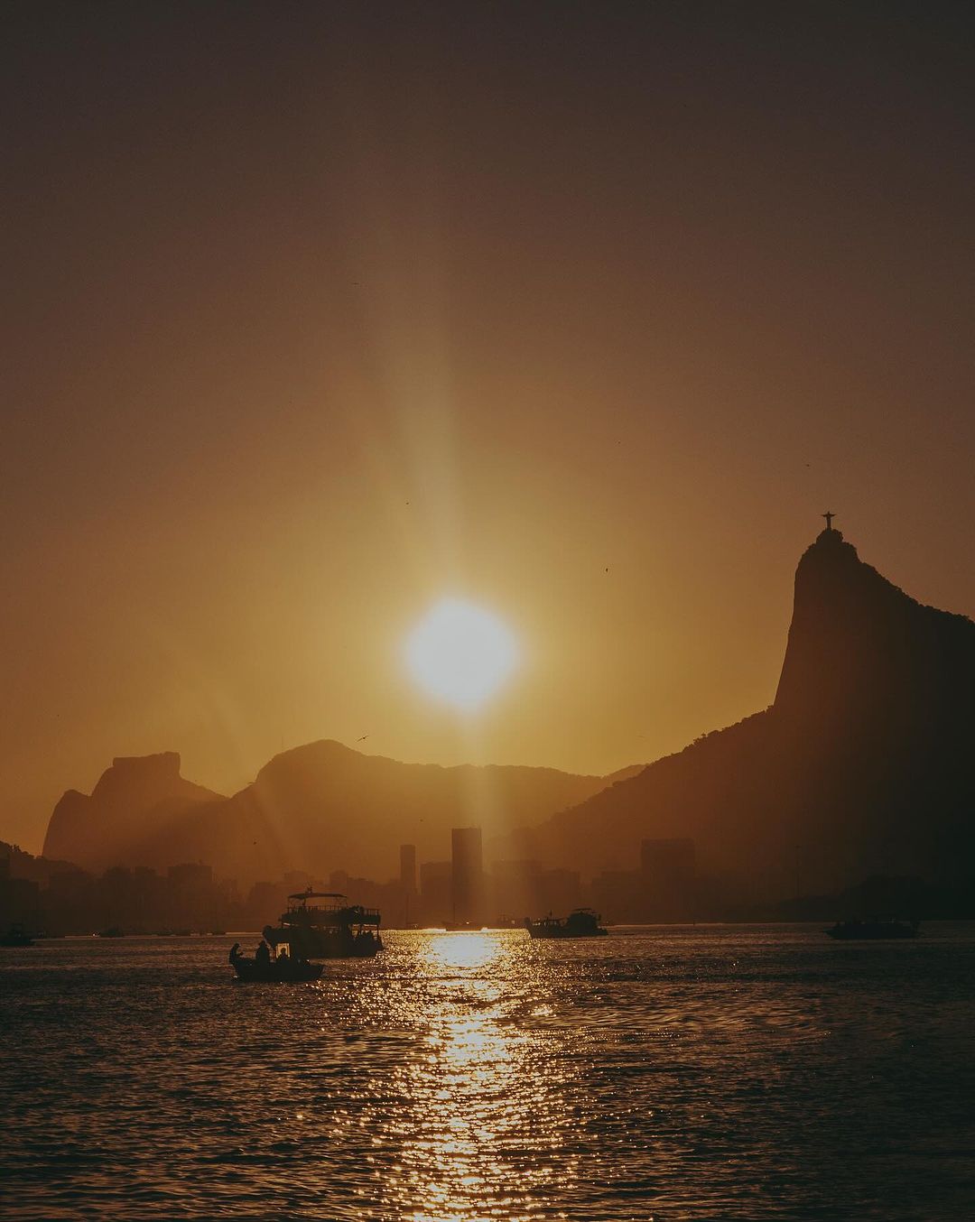 Golden hour view of Rio de Janeiro with mountains, city skyline, and boats on the water under a bright sun and sky.