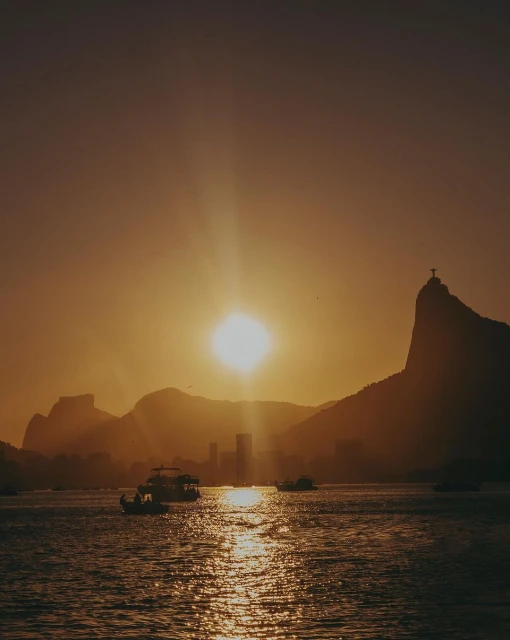 Golden hour view of Rio de Janeiro with mountains, city skyline, and boats on the water under a bright sun and sky.