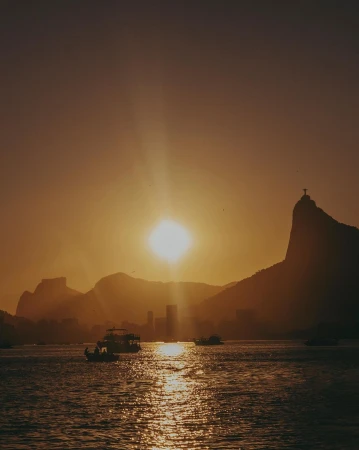 Golden hour view of Rio de Janeiro with mountains, city skyline, and boats on the water under a bright sun and sky.