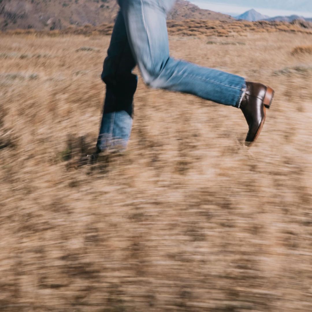 Person running across dry grassy field wearing blue jeans and brown leather boots. Low angle, motion blur and mountains in the background.