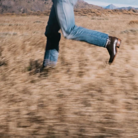 Person running across dry grassy field wearing blue jeans and brown leather boots. Low angle, motion blur and mountains in the background.