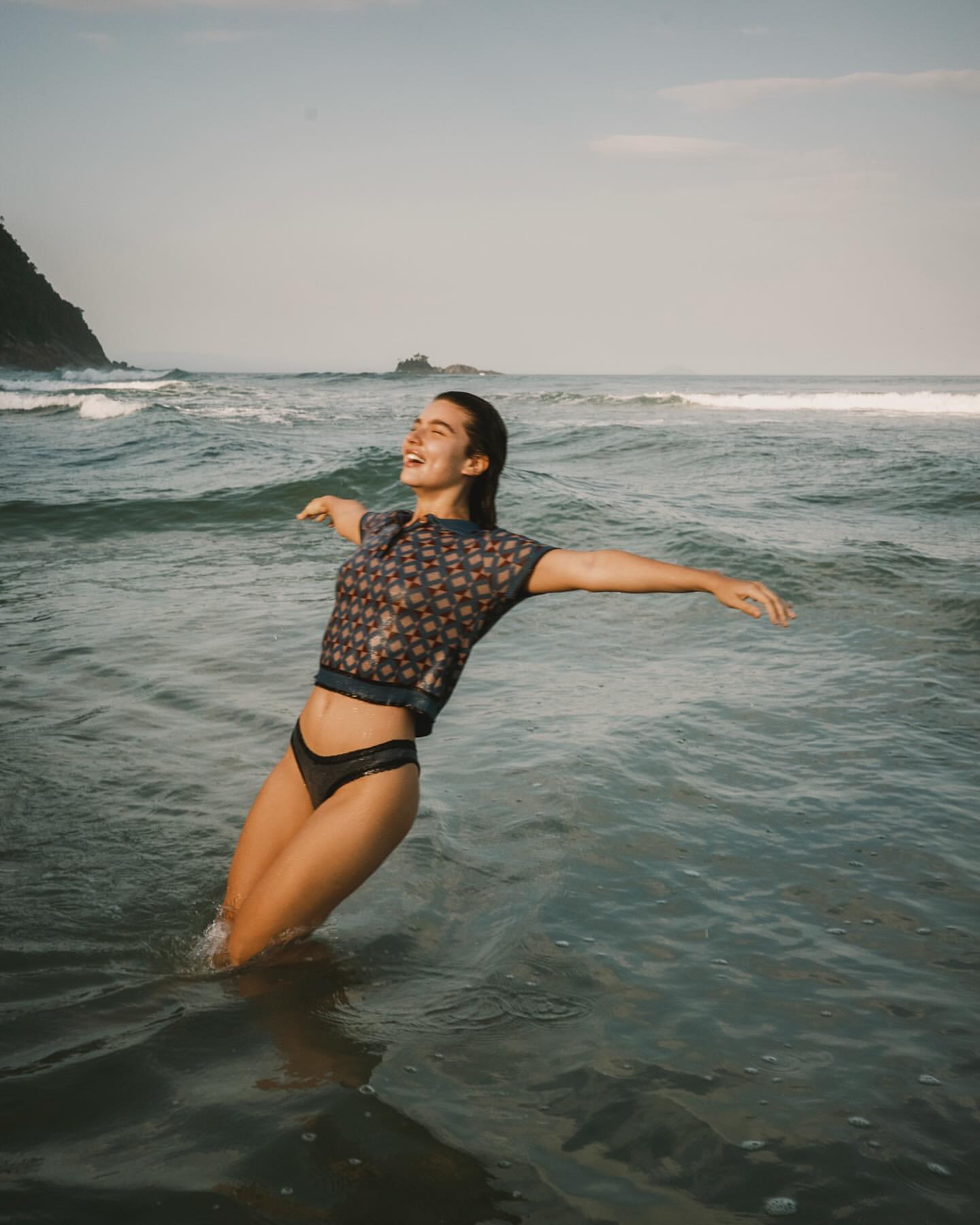 A woman stands waist-deep in the ocean, arms outstretched, smiling against a backdrop of gentle waves and a distant, rocky coastline.