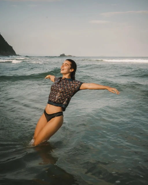 A woman stands waist-deep in the ocean, arms outstretched, smiling against a backdrop of gentle waves and a distant, rocky coastline.