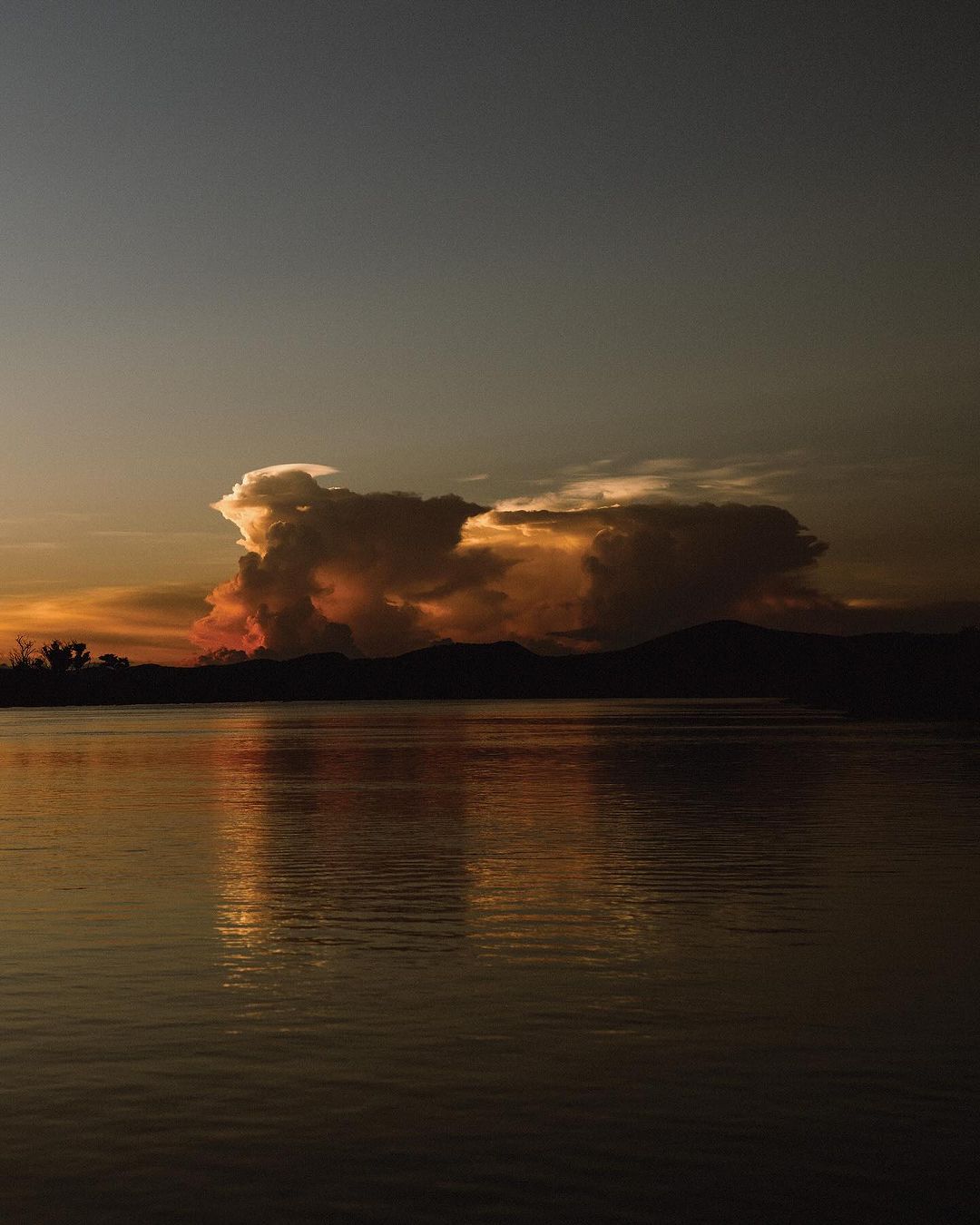 Golden hour illuminates the lake with silhouetted mountains and trees. The sunset clouds add drama and depth to the serene water landscape.