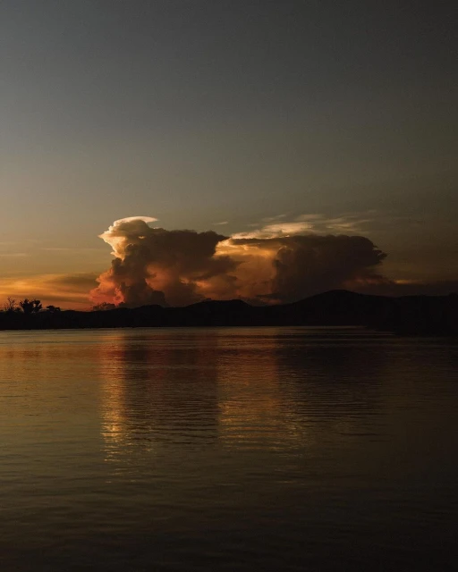 Golden hour illuminates the lake with silhouetted mountains and trees. The sunset clouds add drama and depth to the serene water landscape.