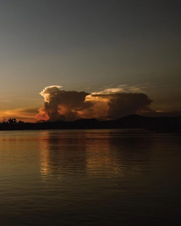 Golden hour illuminates the lake with silhouetted mountains and trees. The sunset clouds add drama and depth to the serene water landscape.