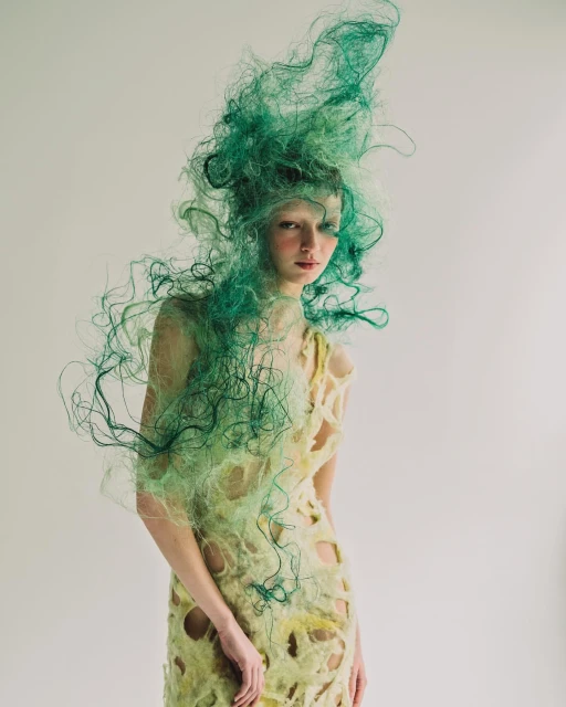 A young woman with green hair extensions and a flowing, textured dress, poses in a fashion editorial shot against a neutral backdrop.