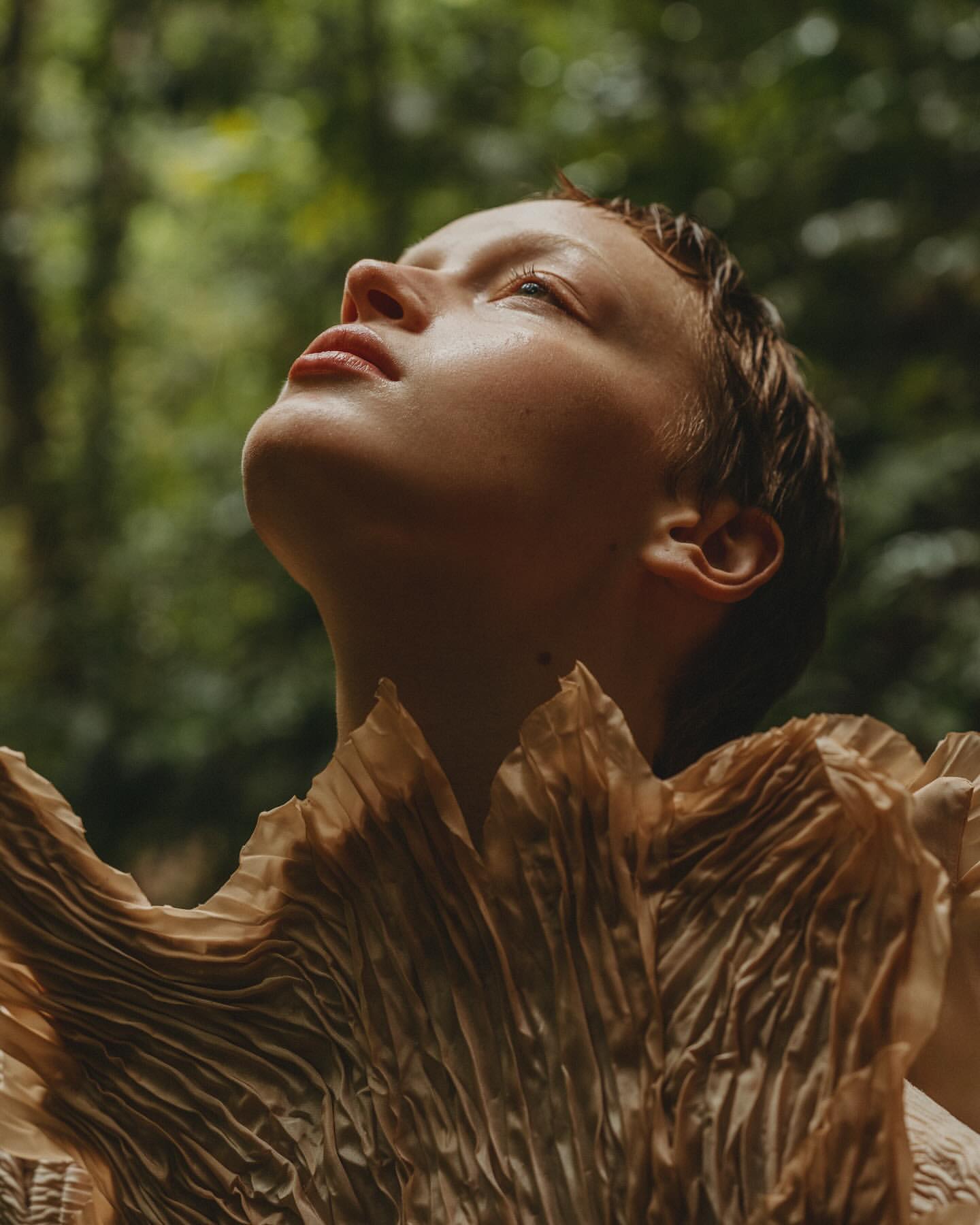 Eye-level portrait of a fair-skinned adult female looking upwards with a leafy green forest background and wearing a textured garment.