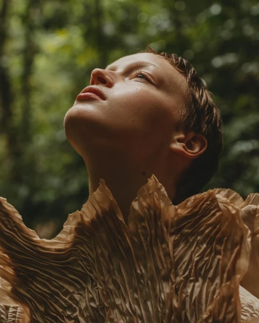 Eye-level portrait of a fair-skinned adult female looking upwards with a leafy green forest background and wearing a textured garment.