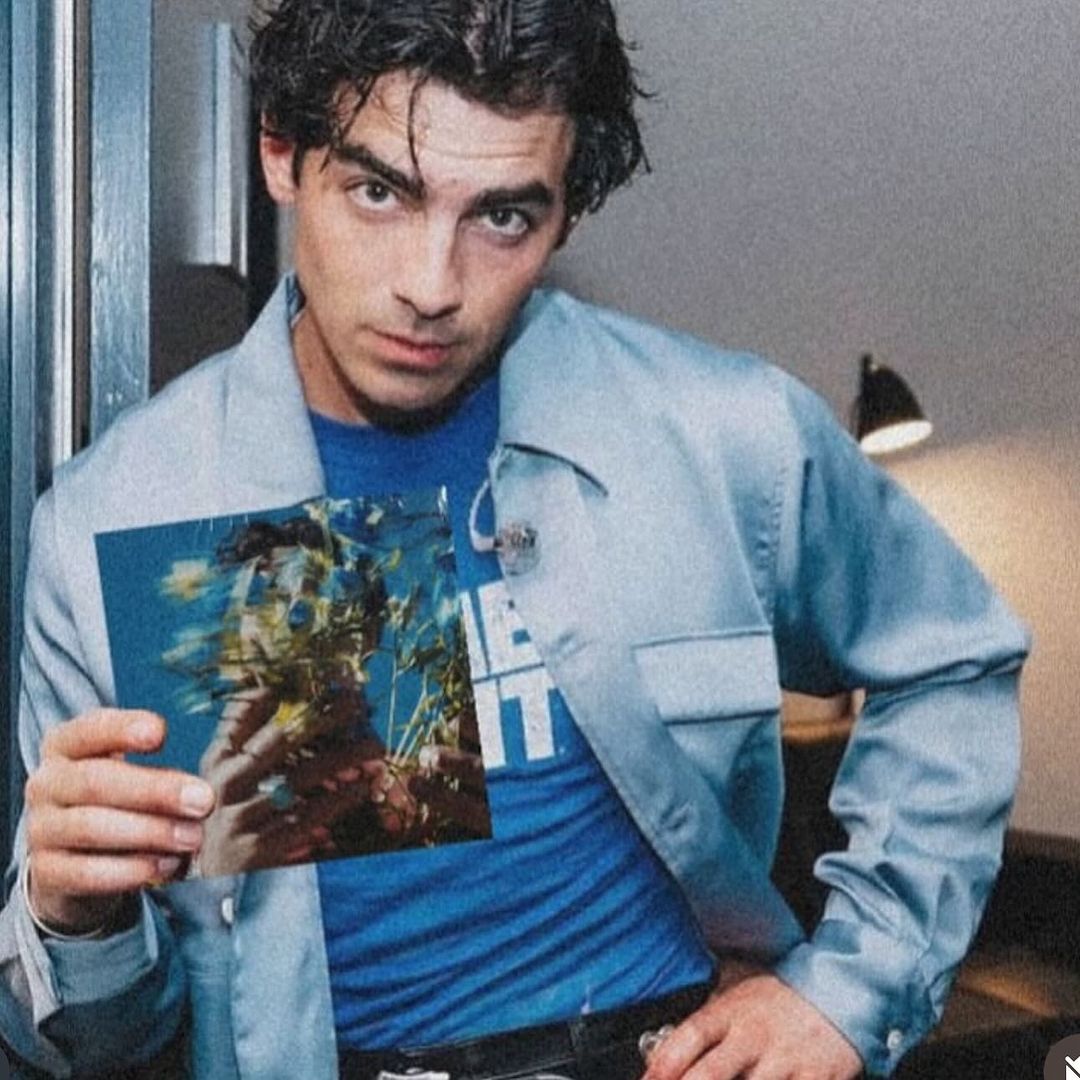Adult man in a blue shirt and blue jacket, holding an object, poses indoors with a dark background and overhead lamp.