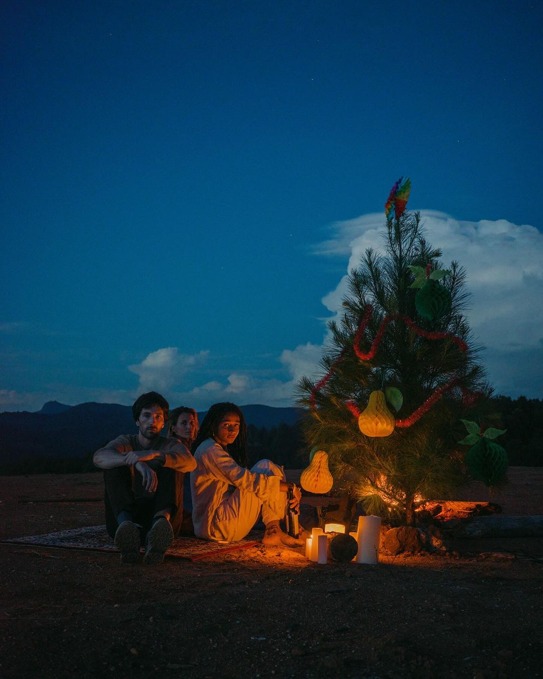 Three people are sitting outside near a decorated Christmas tree lit by candles at night with distant hills under a blue sky.