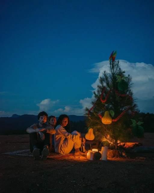 Three people are sitting outside near a decorated Christmas tree lit by candles at night with distant hills under a blue sky.
