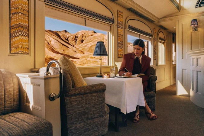 Woman in a blazer writing at a table on a train with a desert view, elegant travel, vintage interior, and refined ambiance.