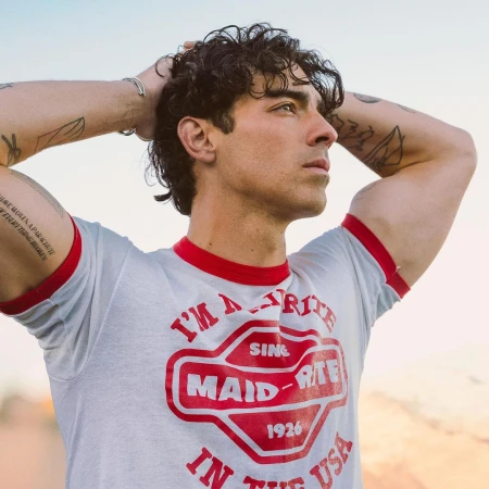Joe Jonas with curly dark hair and tattoos wearing a grey ringer t-shirt with red trim posing outdoors.
