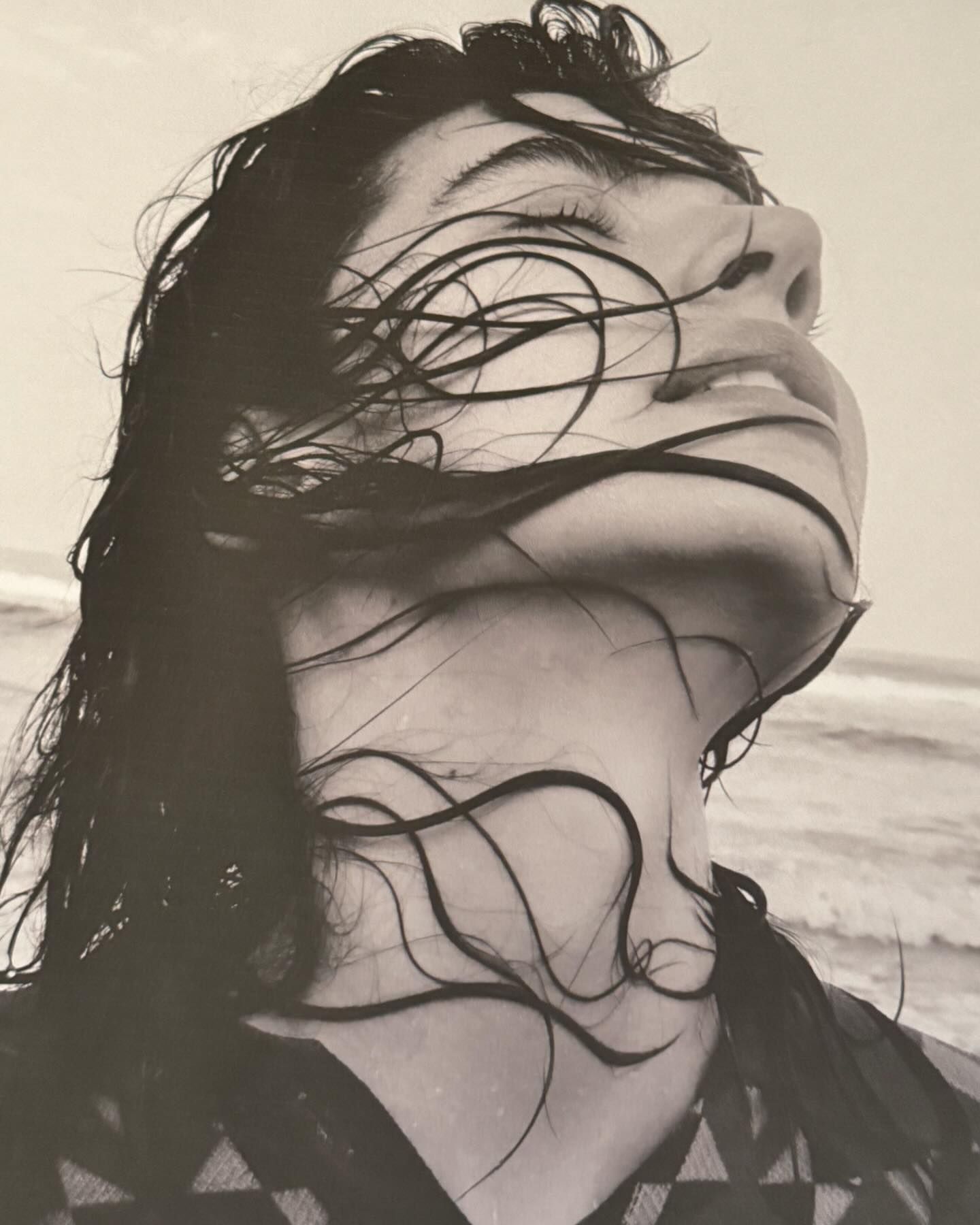 Close-up portrait of a woman with wet dark hair draped across her face, tilting her head back against a hazy beach background.