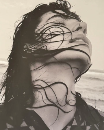 Close-up portrait of a woman with wet dark hair draped across her face, tilting her head back against a hazy beach background.