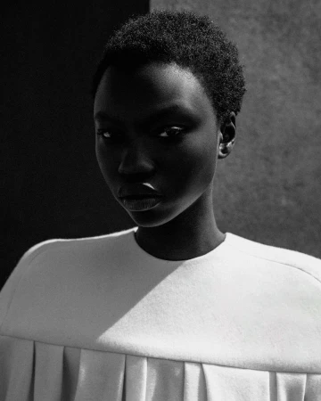Monochrome portrait of a Black woman with short hair wearing a structured white top with pleated detailing under stark directional light.