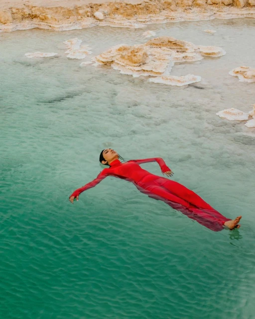 A woman in a red dress floats in teal water, arms outstretched, with rocky formations in the background, creating a serene and picturesque scene.