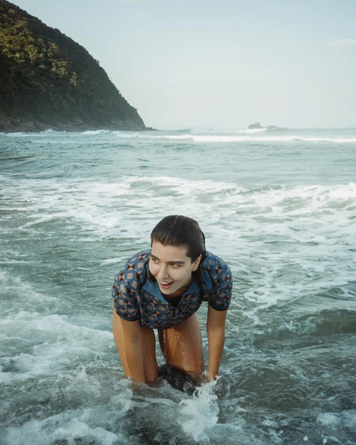 A woman in a patterned top stands in ocean waves, smiling, with a forested hillside in the background under a bright sky.