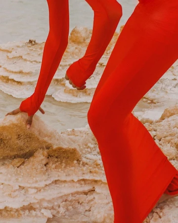 Partial view of a person in a red jumpsuit with long sleeves and flared legs kneeling on a textured, tan and white surface.