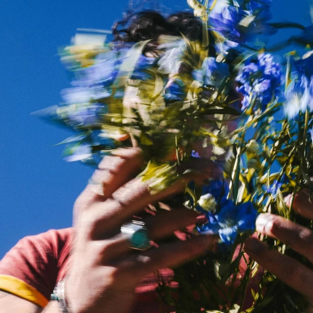 Person with a bouquet of blue flowers held up to their face against a bright blue sky. A ring is visible on the person's finger.