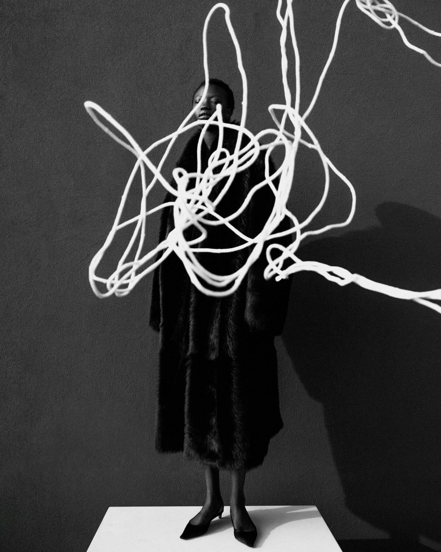 Portrait of a Black woman in a fur coat standing on a pedestal surrounded by white string against a grey backdrop in black and white.