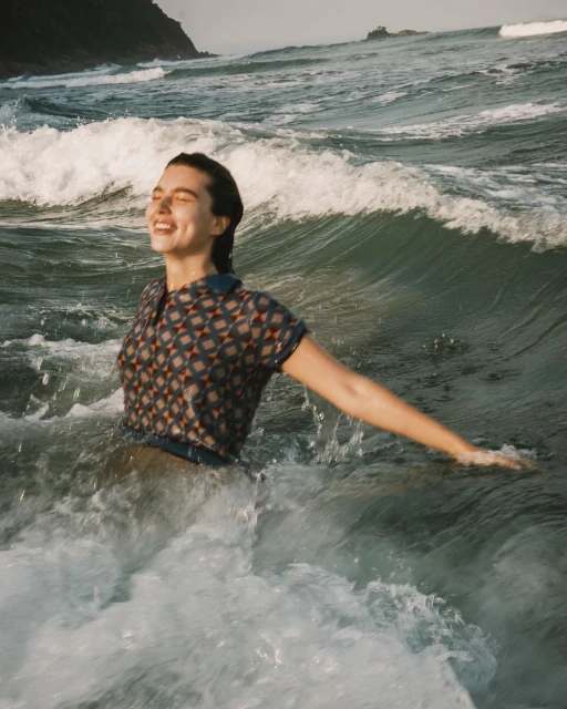 Woman smiling and wading in ocean waves with sunlit water and a distant rocky coastline, enjoying a sunny day at the beach.