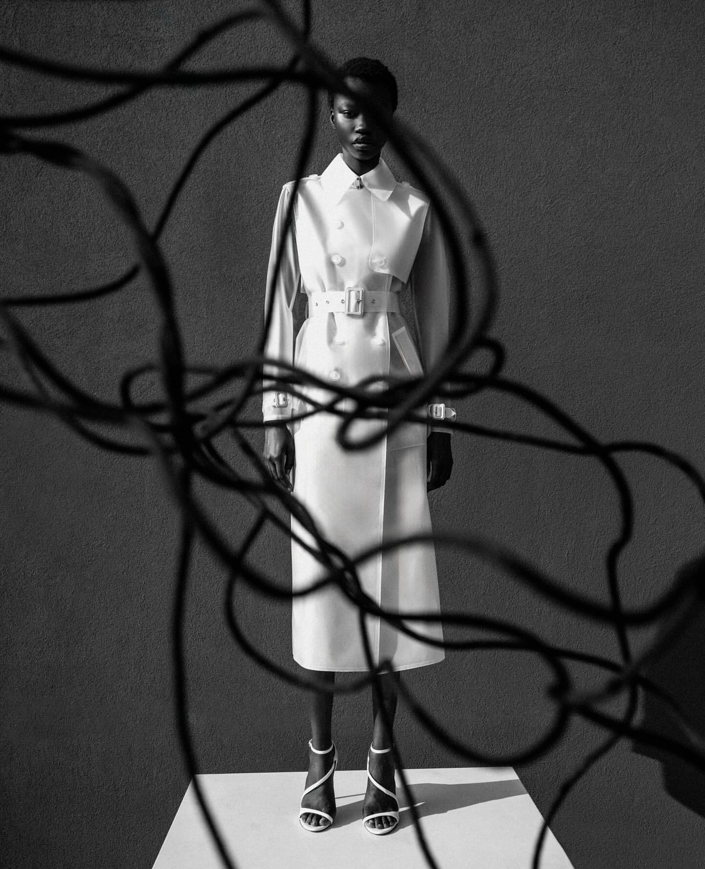 A Black female model stands in a studio wearing a double-breasted trench coat, strap heels, and posing with abstract tangled wires.