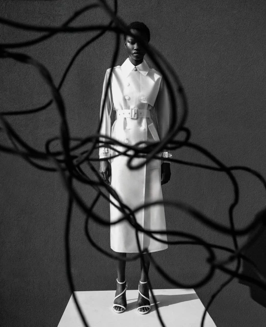 A Black female model stands in a studio wearing a double-breasted trench coat, strap heels, and posing with abstract tangled wires.
