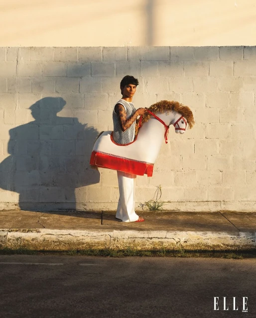 A person with tattoos wearing a horse costume in an outdoor setting, posing in front of a white brick wall on a sunny day.
