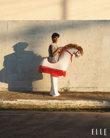 A person with tattoos wearing a horse costume in an outdoor setting, posing in front of a white brick wall on a sunny day.