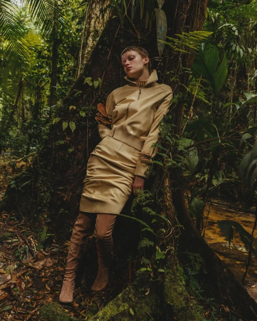 A slender, blonde woman in a gold skirt suit and thigh-high boots leans against a large tree in a verdant jungle environment.