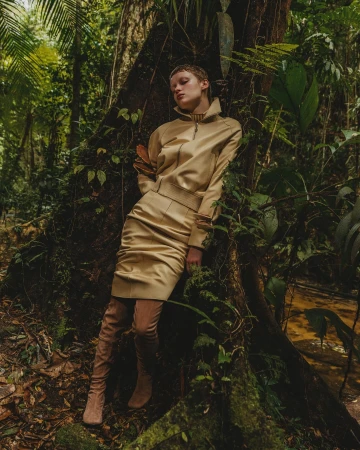 A slender, blonde woman in a gold skirt suit and thigh-high boots leans against a large tree in a verdant jungle environment.