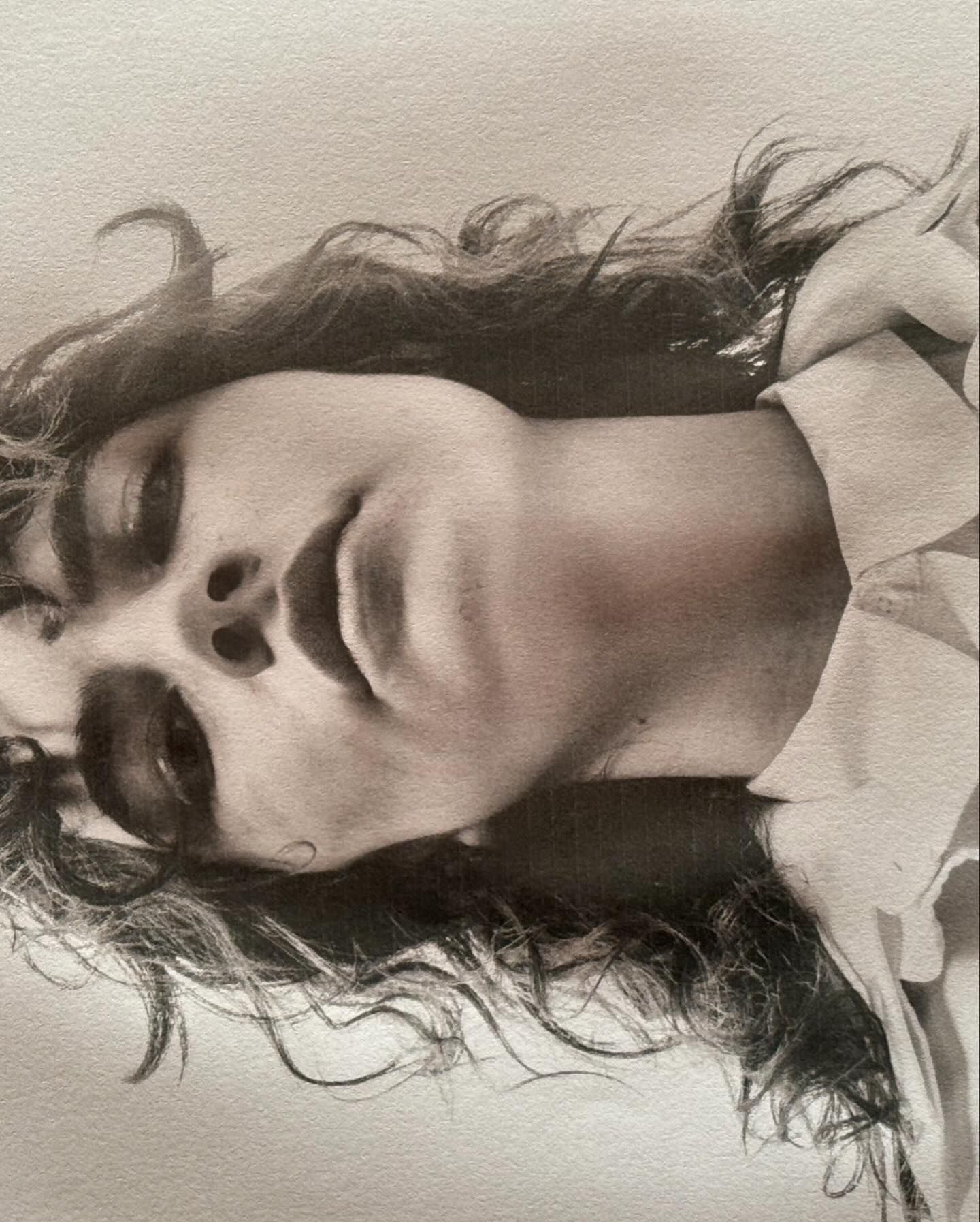 Black and white portrait of a person with curly hair looking up toward the camera wearing a white collared shirt against a blank background.