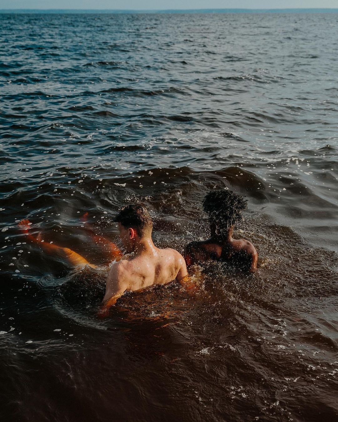 Two shirtless men, one light-skinned and one dark-skinned, stand shoulder-to-shoulder waist-deep in the ocean, facing away from the camera.