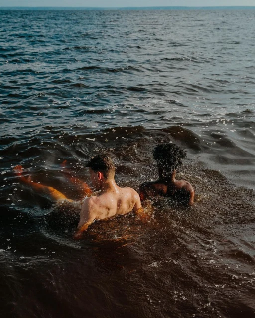 Two shirtless men, one light-skinned and one dark-skinned, stand shoulder-to-shoulder waist-deep in the ocean, facing away from the camera.