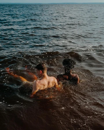 Two shirtless men, one light-skinned and one dark-skinned, stand shoulder-to-shoulder waist-deep in the ocean, facing away from the camera.