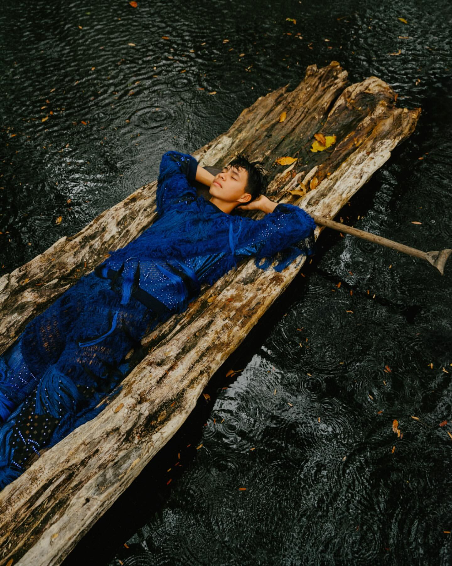 A young adult man in a blue outfit lies on a weathered wooden raft in a pool of water with an oar nearby in an outdoor setting.