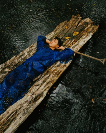 A young adult man in a blue outfit lies on a weathered wooden raft in a pool of water with an oar nearby in an outdoor setting.