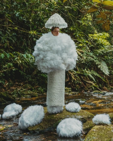 Person dressed in a white mushroom costume stands in a lush green forest next to a stream in a surreal, whimsical portrait. 
