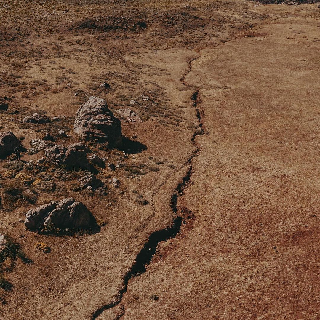 Aerial view of a dry, grassy landscape with rock formations and a deep crack running through the terrain, creating a textured, earthy scene.