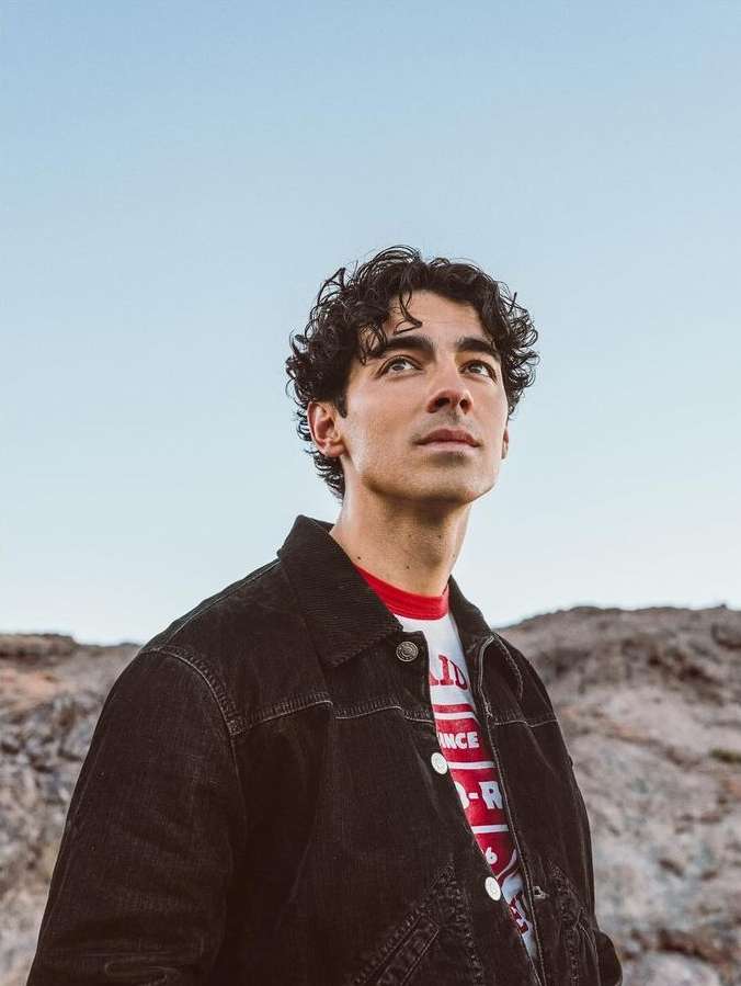 Portrait of a handsome, curly-haired adult man in a black denim jacket looking up towards the sky in a rocky, outdoor setting.