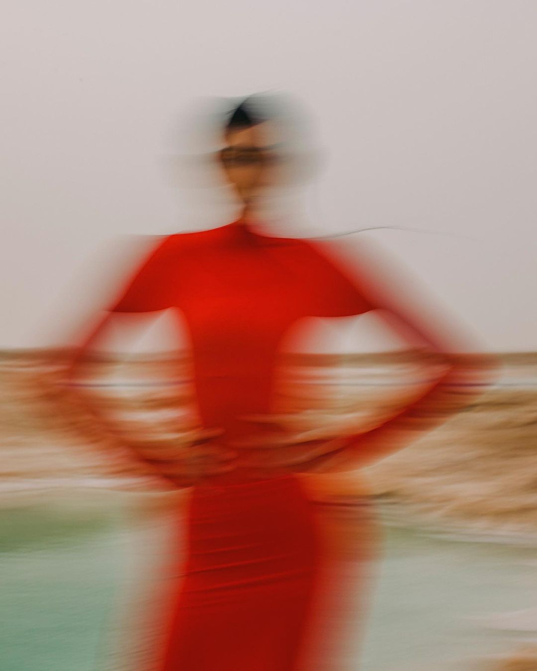 Woman with motion blur wears red dress posing on a sandy surface near water with a light sky background, creating a dreamy and artistic effect.