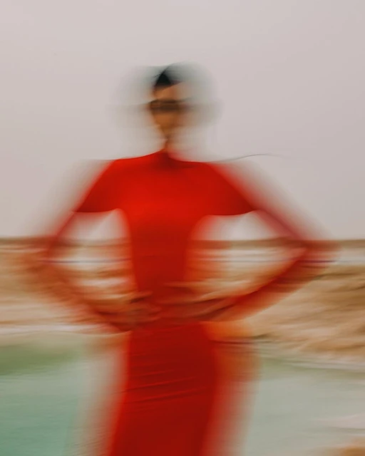 Woman with motion blur wears red dress posing on a sandy surface near water with a light sky background, creating a dreamy and artistic effect.
