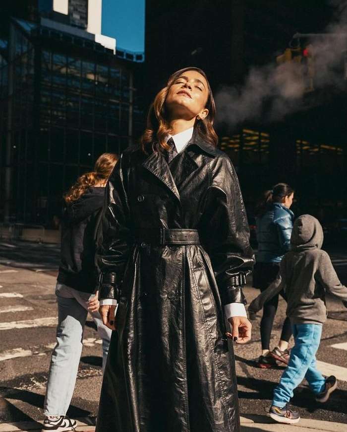 Woman in a black trench coat standing on a city street with pedestrians and buildings in the background, bathed in warm light.