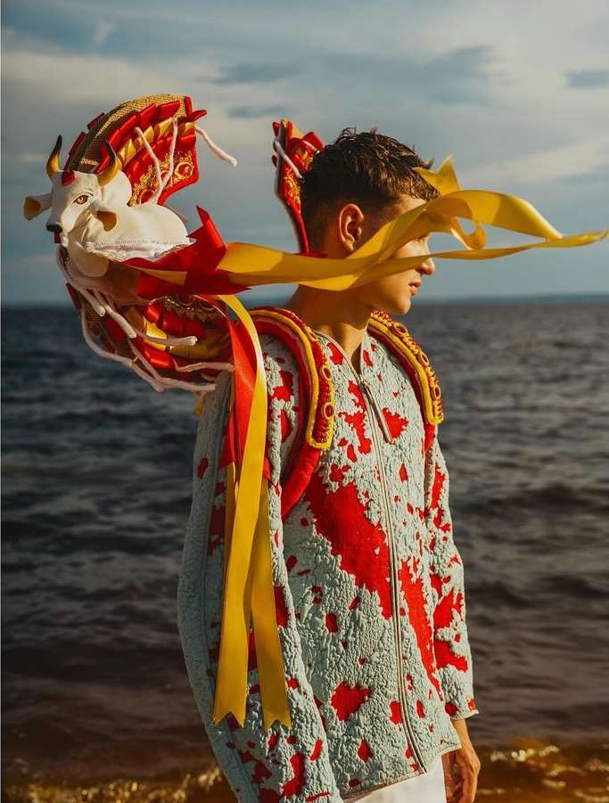 An individual stands near a shore wearing a jacket with red detailing and a decorative white bull headpiece with yellow ribbons, ocean backdrop.